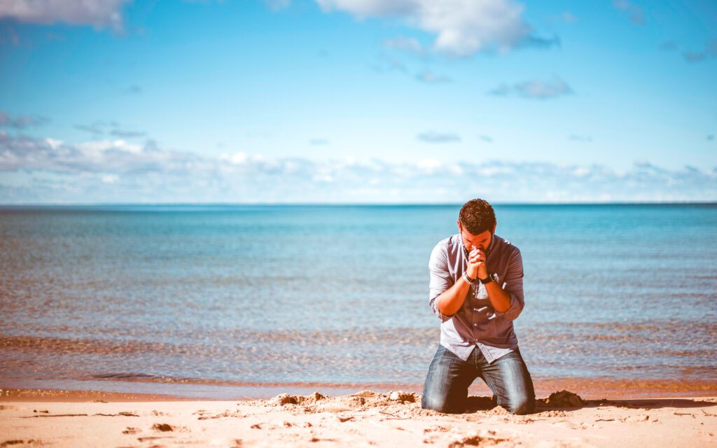 A man kneels on a sandy beach with his face between his hands. He seems to be praying. The sky is partly cloudy and the waters are calm.